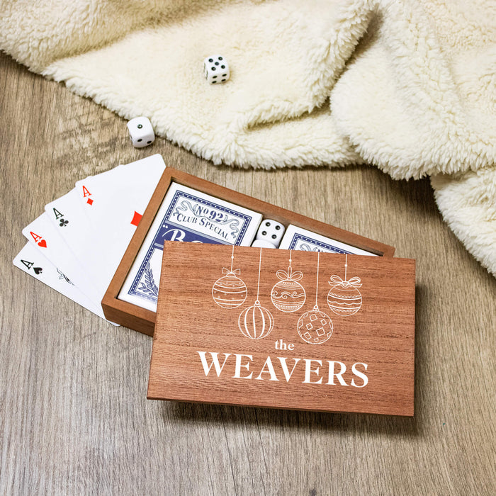 A wooden gift box printed with the words 'THE WEAVERS' and white ornament illustrations sits open on a wood surface, with playing cards and dice visible inside and around it. A cream plush blanket is draped in the background.