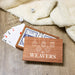 A wooden gift box printed with the words 'THE WEAVERS' and white ornament illustrations sits open on a wood surface, with playing cards and dice visible inside and around it. A cream plush blanket is draped in the background.