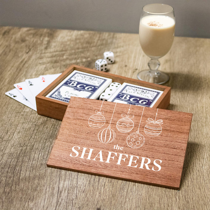 A wooden gift box containing Playing Cards, with a matching printed wooden lid in front that reads 'THE SHAFFERS' and shows white line drawings of hanging ornaments. The box sits on a wooden table next to playing cards, dice, and a glass of milk.