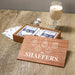 A wooden gift box containing Playing Cards, with a matching printed wooden lid in front that reads 'THE SHAFFERS' and shows white line drawings of hanging ornaments. The box sits on a wooden table next to playing cards, dice, and a glass of milk.