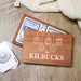 A personalized wooden gift box set for a deck of cards, featuring a front cover printed with 'THE KILBUCKS' and white hanging ornament illustrations. The box is shown open with blue-backed cards and white dice inside, alongside a deck of cards and a die on a rustic wood surface with a cream blanket in the background.