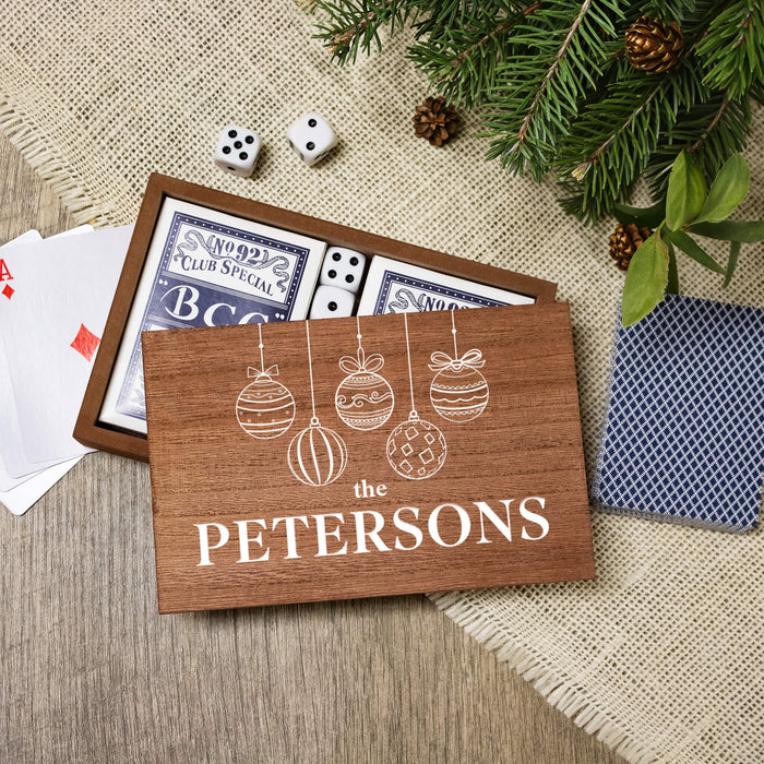 A personalized wooden gift box set for dominoes, featuring a custom printed front lid with white text reading 'THE PETERSONS' and white line art of hanging ornaments. Inside, the dominoes and playing cards are visible.