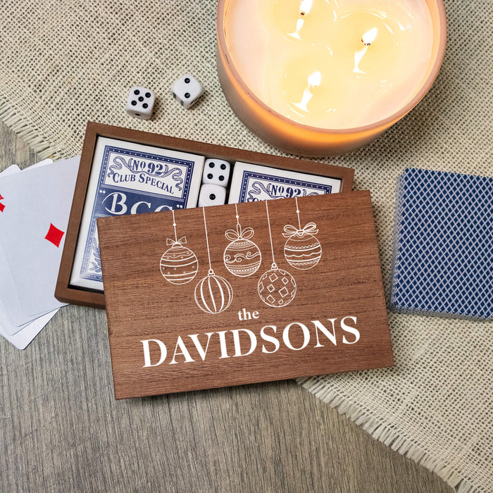 A personalized wooden playing card box printed with 'THE DAVIDSONS' and white ornament illustrations, shown on a burlap surface with playing cards, dice, a lit candle, and a blue patterned card nearby.