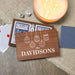 A personalized wooden playing card box printed with 'THE DAVIDSONS' and white ornament illustrations, shown on a burlap surface with playing cards, dice, a lit candle, and a blue patterned card nearby.