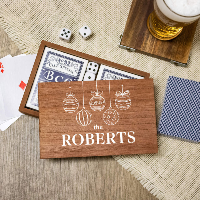 A wooden personalized playing card box with white printed Christmas ornament illustrations and the name 'THE ROBERTS' in bold letters, shown open with playing cards inside. The box is styled on a woven mat with playing cards, dice, and a glass of beer nearby.