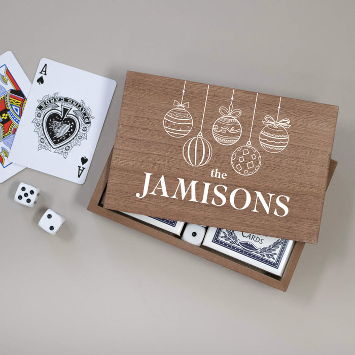 A wooden gift box with a custom printed lid reading 'the Jamisons' and decorated with white hanging ornament illustrations. Inside the box are playing cards, with an Ace of Spades card and two white dice placed beside it on a neutral gray background.