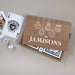 A wooden gift box with a custom printed lid reading 'the Jamisons' and decorated with white hanging ornament illustrations. Inside the box are playing cards, with an Ace of Spades card and two white dice placed beside it on a neutral gray background.