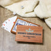 A personalized wooden Christmas card game set on a wood floor with a cream sherpa blanket. The set includes a wooden box printed with 'MERRY CHRISTMAS' THE JONES FAMILY 2026 and a snowflake, containing playing cards and dice.