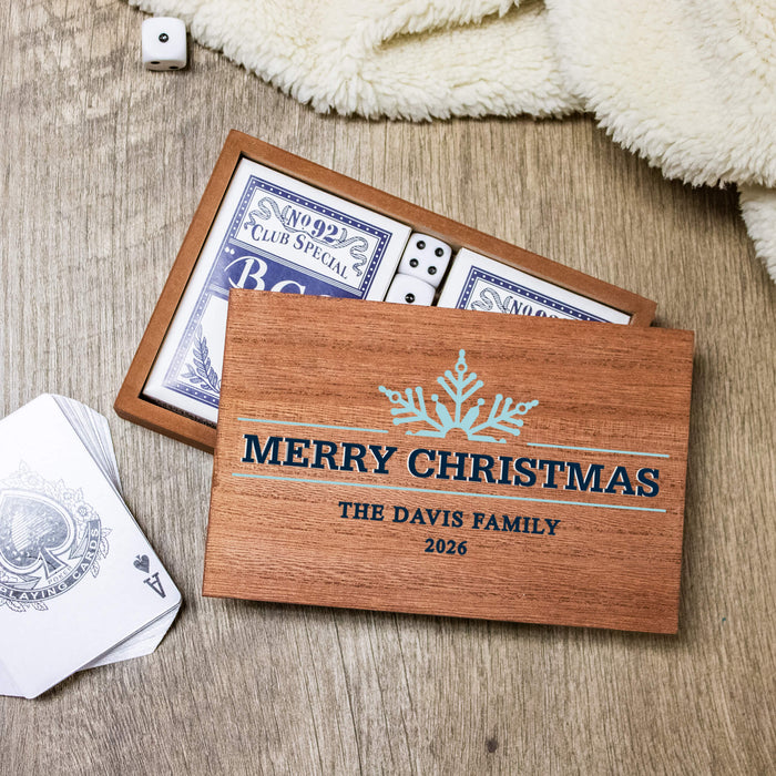 A personalized wooden Christmas gift box with a printed snowflake and the text 'MERRY CHRISTMAS' THE DAVIS FAMILY 2026, shown open with playing cards and dice on a wood surface with a cream blanket nearby.
