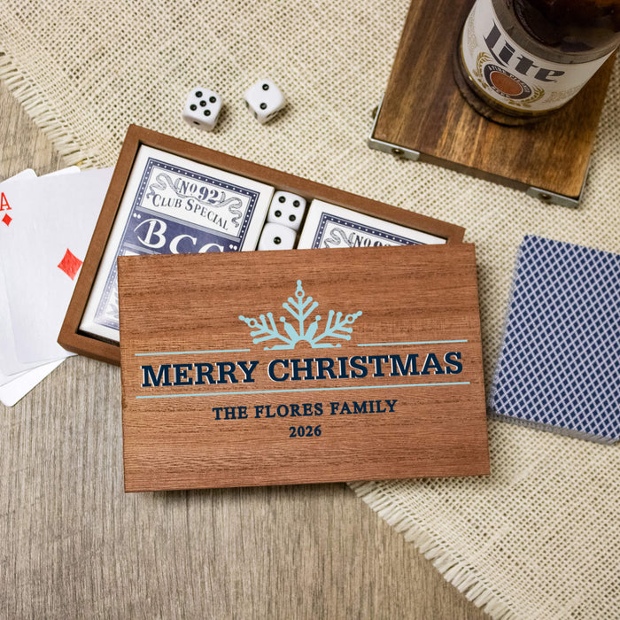 A personalized wooden Christmas-themed card game box set on a rustic tabletop. The set includes a wooden box printed with 'MERRY CHRISTMAS' THE FLORES FAMILY 2026 and a snowflake design, containing a deck of playing cards with blue and white backs, dice, playing cards, and a blue patterned card on a woven mat.