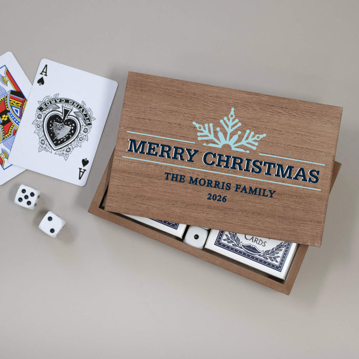 A personalized wooden Christmas card game box with a hinged lid printed with 'MERRY CHRISTMAS' THE MORRIS FAMILY 2026 and a snowflake design, shown open with playing cards and dice on a neutral gray background.