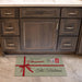 A rectangular holiday-themed doormat placed in front of dark wood bathroom cabinets. The mat is light sage green with white polka dots, a red ribbon graphic, and the text 'Merry Christmas' and 'The Halsters' with small postage-style holiday stamps.