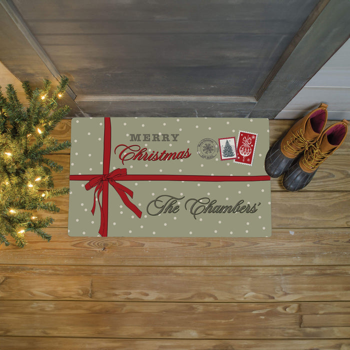 A personalized holiday doormat styled on a wooden porch floor next to a Christmas tree and a pair of boots. The doormat is green with white polka dots, featuring a red ribbon graphic, 'Merry Christmas' text, and 'The Chambers' printed in dark green script.