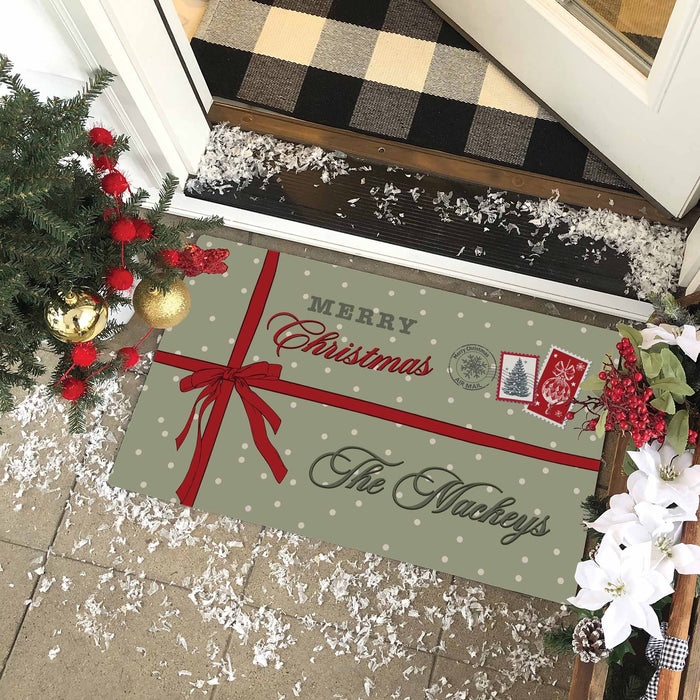 A personalized Christmas doormat styled on a tiled entryway floor, decorated with holiday garlands and fake snow. The mat is sage green with white polka dots, featuring a gift box graphic with a red ribbon, the text 'MERRY Christmas The Mackeys', and small red and gray holiday stamps.