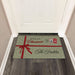 A rectangular holiday doormat placed in front of a white door on a dark wood floor. The mat is light sage green with white polka dots, a red ribbon graphic, and the text 'Merry Christmas' and 'The Finkles' with small festive illustrations.