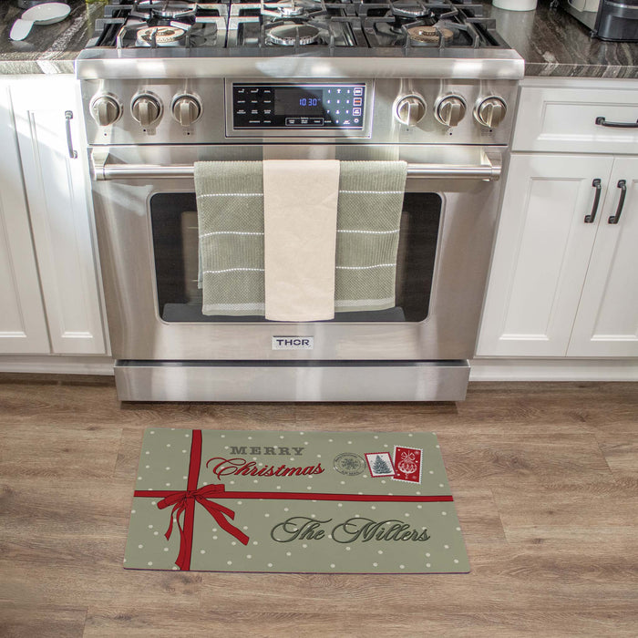 A rectangular kitchen floor mat styled like a wrapped Christmas gift, featuring the text 'Merry Christmas The Millers' and postage-style stamps, placed in front of a stainless steel THOR gas range on a wood floor.