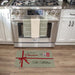 A rectangular kitchen floor mat styled like a wrapped Christmas gift, featuring the text 'Merry Christmas The Millers' and postage-style stamps, placed in front of a stainless steel THOR gas range on a wood floor.
