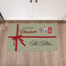A rectangular holiday doormat with a sage green background, white polka dots, a red ribbon graphic, and the text 'Merry Christmas' and 'The Tellers', shown placed on a wood floor in front of white cabinet doors.
