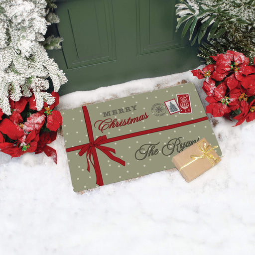 A personalized Christmas doormat in sage green with white polka dots, a red ribbon, and 'Merry Christmas The Ryans' printed on it, styled in a snowy porch setting next to poinsettias and a green door.