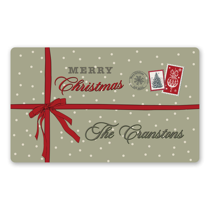 A rectangular holiday doormat with a sage green background, white polka dots, a red ribbon bow across the center, and the text 'MERRY Christmas' and 'The Cranstons,' plus small postage-style Christmas illustrations.