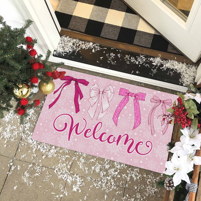 A pink rectangular doormat with decorative bow illustrations and the word 'Welcome' in script, placed at a front door threshold. The scene includes holiday decorations, a black-and-white plaid rug, and snow on the porch.