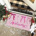 A pink rectangular doormat with decorative bow illustrations and the word 'Welcome' in script, placed at a front door threshold. The scene includes holiday decorations, a black-and-white plaid rug, and snow on the porch.