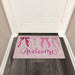 A pink welcome doormat with decorative bows and the word 'Welcome' in script, placed in front of a white door on a dark wood floor.