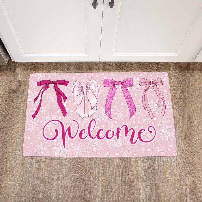 A rectangular pink welcome doormat with four illustrated bows in shades of pink and white, plus the word 'Welcome' in cursive, placed on a wood floor in front of white cabinet doors.