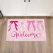 A rectangular pink welcome doormat with four illustrated bows in shades of pink and white, plus the word 'Welcome' in cursive, placed on a wood floor in front of white cabinet doors.