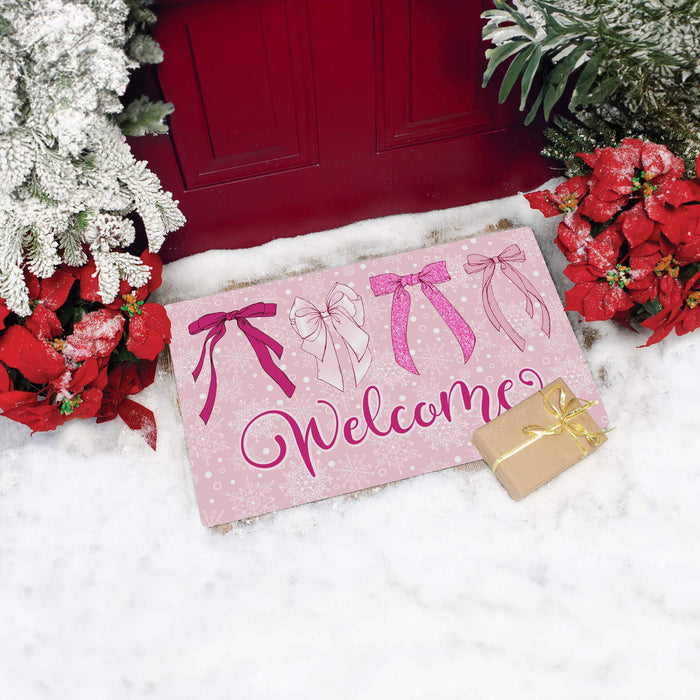 A pink holiday doormat with the word 'Welcome' in cursive and four bow illustrations, styled on white snow with a red door, poinsettias, and a small wrapped gift box.