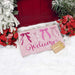 A pink holiday doormat with the word 'Welcome' in cursive and four bow illustrations, styled on white snow with a red door, poinsettias, and a small wrapped gift box.