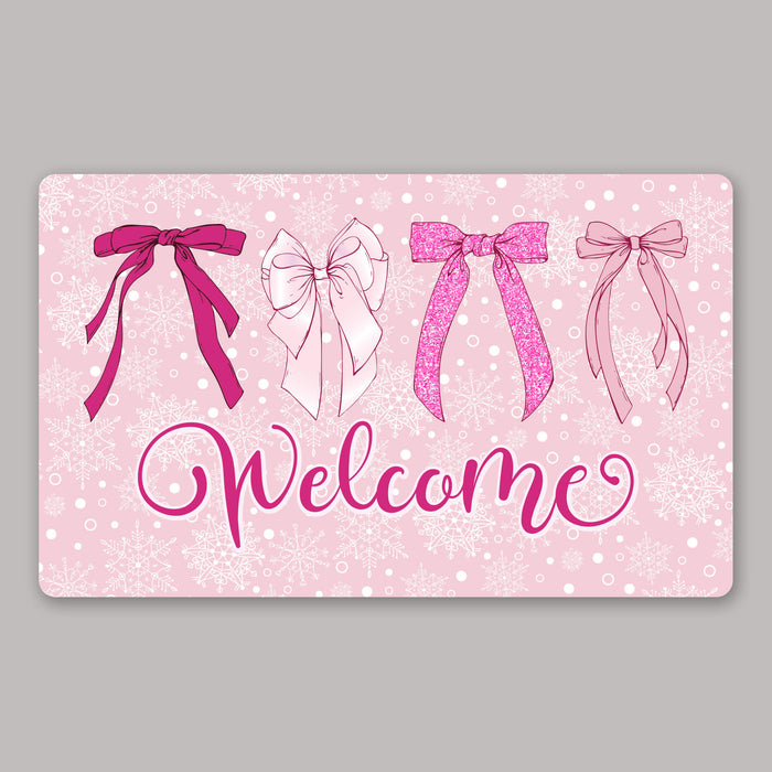 A rectangular pink holiday-themed mat with snowflake patterns, four illustrated ribbon bows in pink tones, and the word 'Welcome' in large cursive script, shown on a plain gray background.