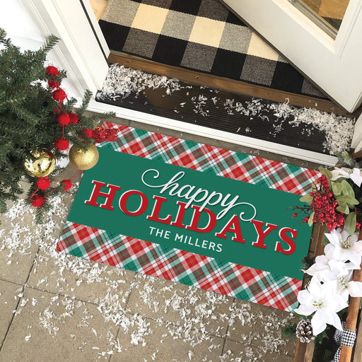 A rectangular holiday doormat with a green center panel reading 'happy HOLIDAYS THE MILLERS' in white and red lettering, bordered by a red, green, and white plaid pattern. The mat is shown on a tiled porch at a doorway with Christmas decorations, a black-and-beige buffalo plaid doormat above, and white faux snow scattered around.