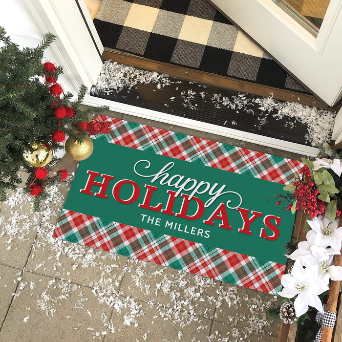 A rectangular holiday doormat with a green center panel reading 'happy HOLIDAYS THE MILLERS' in white and red lettering, bordered by a red, green, and white plaid pattern. The mat is shown on a tiled porch at a doorway with Christmas decorations, a black-and-beige buffalo plaid doormat above, and white faux snow scattered around.