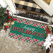 A rectangular holiday doormat with a green center panel reading 'happy HOLIDAYS THE MILLERS' in white and red lettering, bordered by a red, green, and white plaid pattern. The mat is shown on a tiled porch at a doorway with Christmas decorations, a black-and-beige buffalo plaid doormat above, and white faux snow scattered around.
