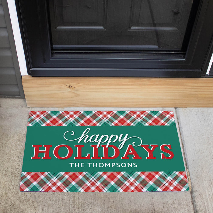 A personalized holiday doormat placed in front of a black front door on a concrete porch. The mat features a green center with the words 'happy HOLIDAYS' and the name 'THE THOMPSONS' in white, bordered by red, green, and white plaid patterns at the top and bottom.
