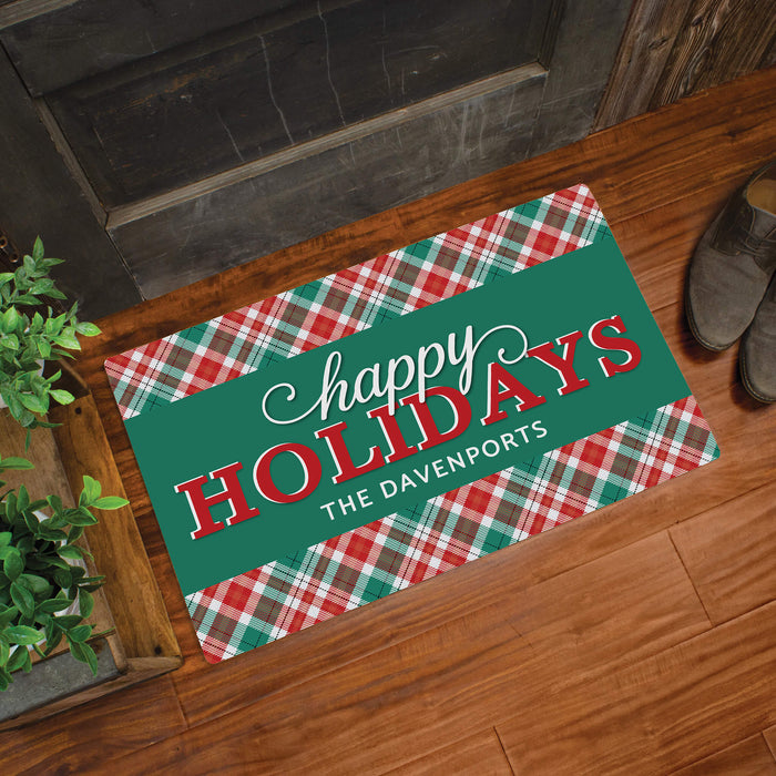 A rectangular holiday doormat with a green center panel reading “happy Holidays” and “THE DAVENPORTS,” surrounded by red, green, and white plaid borders, shown on a wooden floor near a door with shoes and a plant nearby.