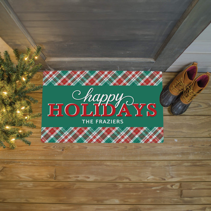 A rectangular holiday doormat with a green center panel reading 'happy HOLIDAYS THE FRAZIER'S' in white and red lettering, bordered by red, green, and white plaid patterns. The mat is placed on a wooden porch floor beside a lit Christmas tree and a pair of brown boots.