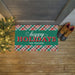 A rectangular holiday doormat with a green center panel reading 'happy HOLIDAYS THE FRAZIER'S' in white and red lettering, bordered by red, green, and white plaid patterns. The mat is placed on a wooden porch floor beside a lit Christmas tree and a pair of brown boots.