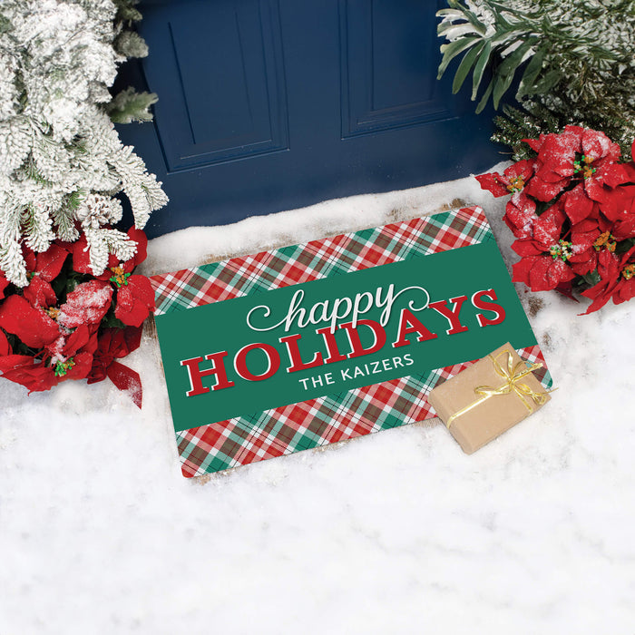 A personalized holiday doormat with a green center panel and red-and-green plaid borders, placed in front of a blue door on white snow with poinsettias and a small wrapped gift nearby.