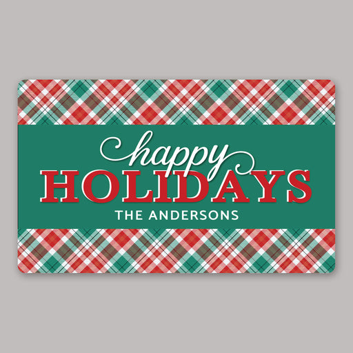 A rectangular holiday door mat with a green center panel reading “happy HOLIDAYS” and “THE ANDERSONS,” framed by red, green, and white plaid borders on a light gray background.