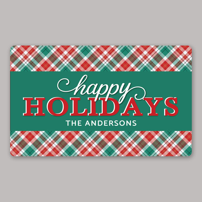 A rectangular holiday door mat with a green center panel reading “happy HOLIDAYS” and “THE ANDERSONS,” framed by red, green, and white plaid borders on a light gray background.