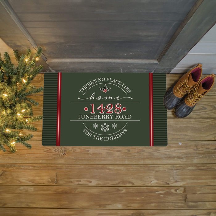 A rectangular holiday doormat with a dark green background and red side stripes, placed on a wooden porch floor. The mat reads “There’s No Place Like Home '1428 Juneberry Road' For The Holidays” with white script and snowflake details.
