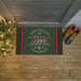 A rectangular holiday doormat with a dark green background and red side stripes, placed on a wooden porch floor. The mat reads “There’s No Place Like Home '1428 Juneberry Road' For The Holidays” with white script and snowflake details.