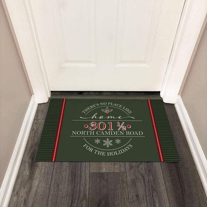 A dark green holiday doormat with red and white text reading 'There's no place like home "2019 NORTH CAMDEN ROAD" FOR THE HOLIDAYS' designed with holly and snowflakes, placed in front of a white door on a wood floor.