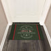 A dark green holiday doormat with red and white text reading 'There's no place like home "2019 NORTH CAMDEN ROAD" FOR THE HOLIDAYS' designed with holly and snowflakes, placed in front of a white door on a wood floor.