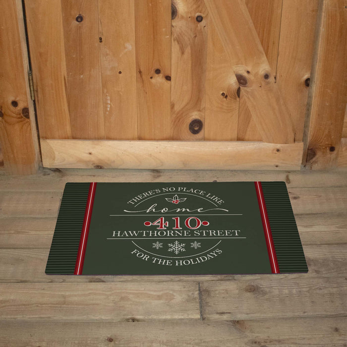A rectangular holiday doormat with a dark green background and red and white printed text reading 'There's no place like home "411 Hawthorne Street" for the holidays,' placed in front of a wooden door and wood floor.