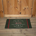 A rectangular holiday doormat with a dark green background and red and white printed text reading 'There's no place like home "411 Hawthorne Street" for the holidays,' placed in front of a wooden door and wood floor.