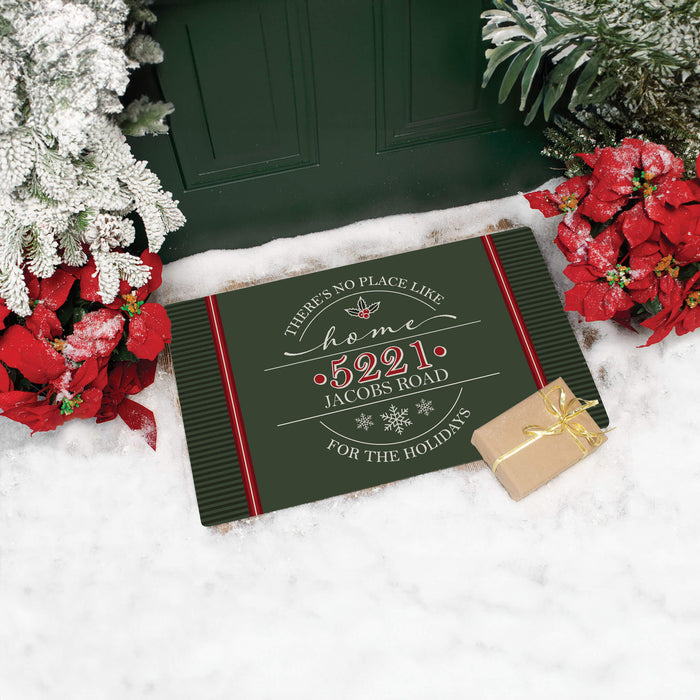 A rectangular holiday doormat with a dark green background, red side borders, and white/red text reading 'There's no place like home "5221 Jacobs Road" for the holidays,' styled on white snow with poinsettias, greenery, and a small wrapped gift box.