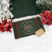 A rectangular holiday doormat with a dark green background, red side borders, and white/red text reading 'There's no place like home "5221 Jacobs Road" for the holidays,' styled on white snow with poinsettias, greenery, and a small wrapped gift box.